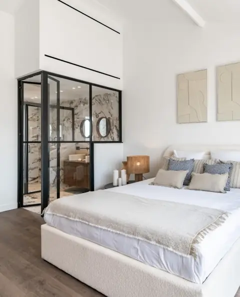 Chambre blanche avec verrière noire donnant sur une salle de bain.