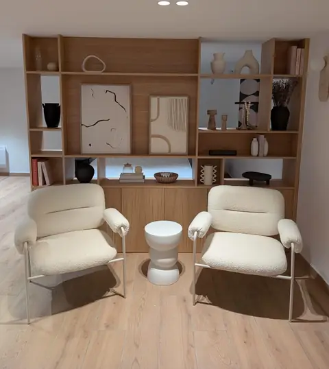 Bibliothèque en chêne derrière deux fauteuils blancs, avec niches et objets décoratifs.