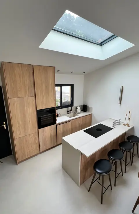 Cuisine sur mesure en bois avec colonnes, îlot blanc et tabourets, sous un puits de lumière.
