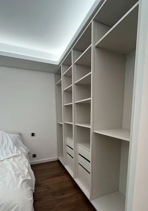 Dressing ouvert blanc avec étagères, tiroirs et éclairage en corniche dans une chambre.