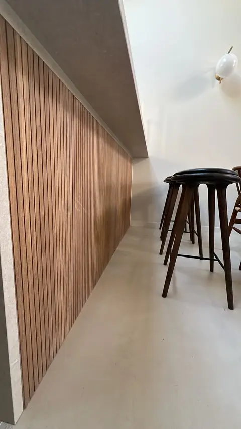 Long mur habillé de tasseaux en bois, avec tabourets de bar au premier plan.