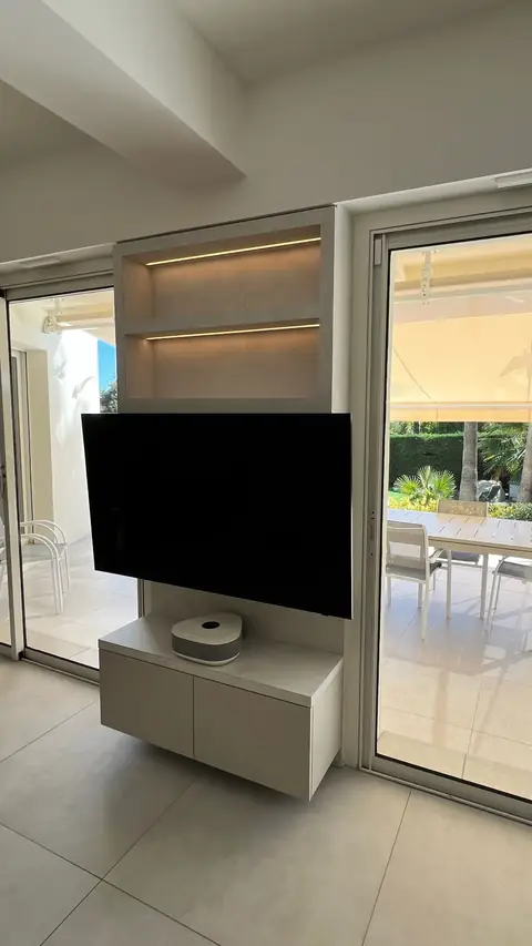 Meuble TV suspendu blanc sous des étagères éclairées, à côté d'une baie vitrée.