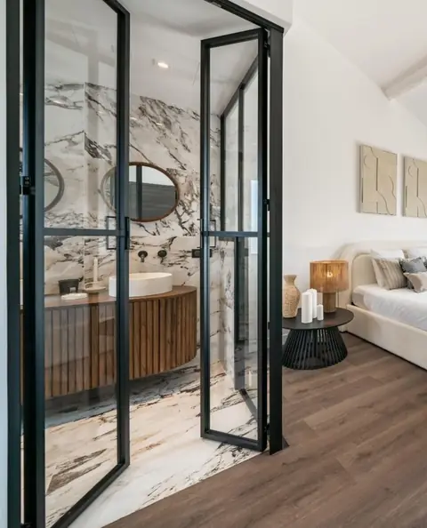 Porte verrière noire ouvrant sur une salle de bain en marbre, depuis une chambre.